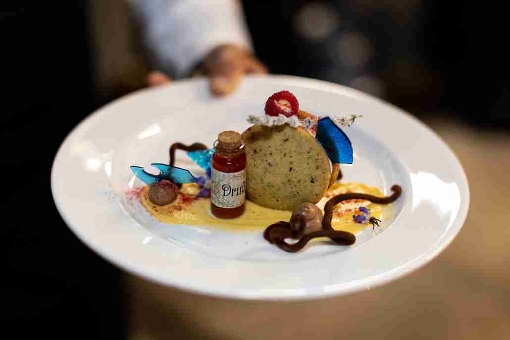 Artfully plated dessert featuring a cake slice, edible flowers, a small bottle labelled "Drink Me," and decorative sauces, showcasing Savoury Chef Catering & Events' commitment to exceptional presentation for corporate events.